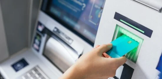 Dłoń wkłada kartę płatniczą do czytnika bankomatu.